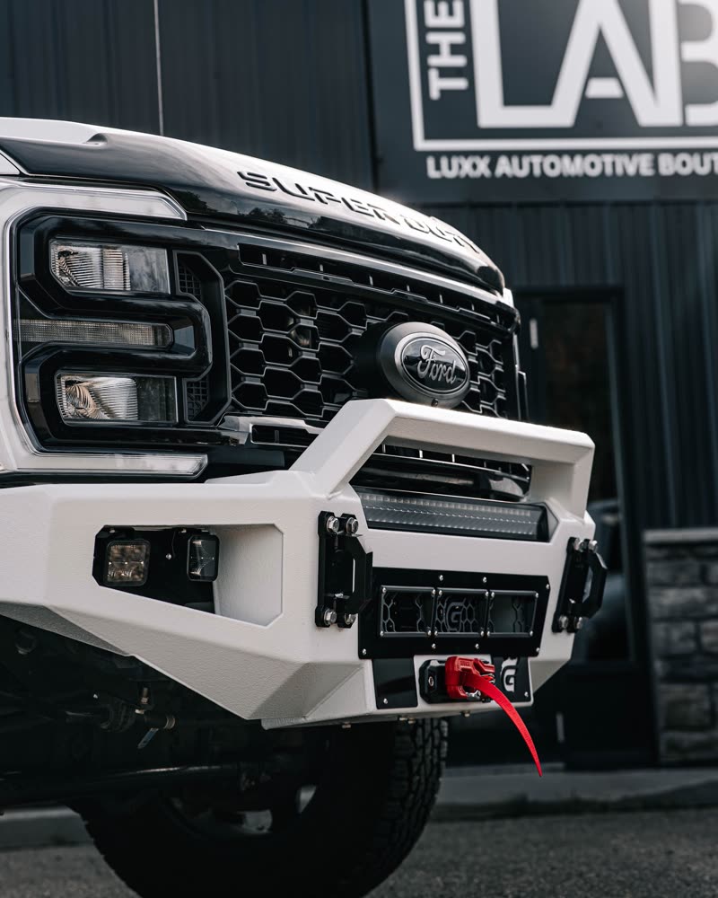 Close-up of a white Gridiron bumper with LED light bar, tow shackles and a red winch hook, with the THE LAB sign behind